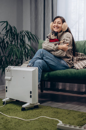 A warmly dressed young female and her dog are warming themselves near an electric radiator.