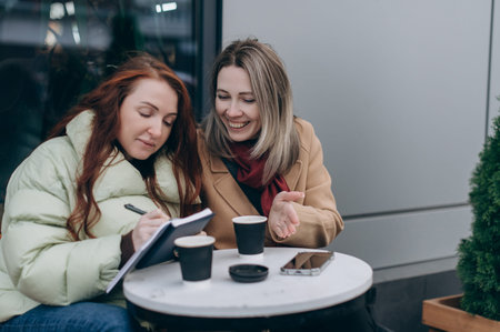 Two cheerful women sitting at an outdoor cafe, writing notes in a notebook and discussing ideas over takeaway coffee during a casual meeting.の写真素材