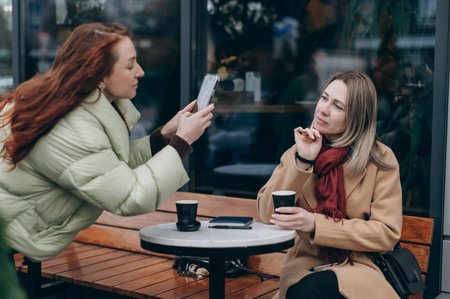 A woman taking a photo of her friend with a smartphone while sitting at an outdoor cafe table and enjoying takeaway coffee in an urban setting.の写真素材