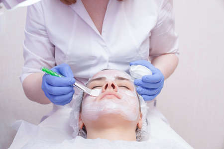 Crop master applying white mask with brush on face and neck of young woman in towel lying on tableの写真素材