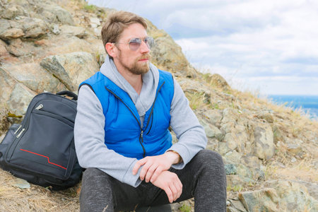 A man in a blue tank top and gray turtleneck stands on a mountain top with a backpack. Close up.