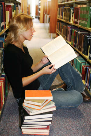 Beautiful young woman sitting on the floor in the library with a stack of books, reading with interestの写真素材