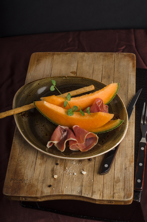 Orange melon with prosciutto and bread sticks close up on wooden texture and black backgroundの写真素材