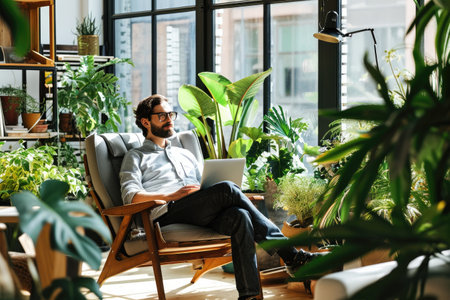 A man dressed in business attire sits in a black chair as he works on a laptop in a modern office, Worker sitting in an eco-friendly office space with plants and daylight, AI Generatedの素材