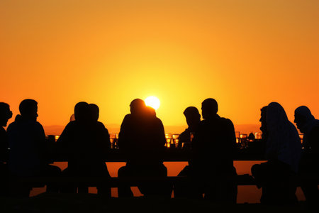 A diverse group of individuals sitting together on a bench, observing the sun as it sets below the horizon, Sunset silhouette of friends at an iftar, AI Generatedの素材