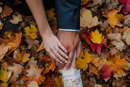 A couple stands together, holding hands, in front of a picturesque pile of autumn leaves, A couple's hands entwined, showing wedding rings against a background of fall leaves, AI Generatedの素材