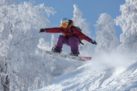 Girl enjoys a thrilling jump on her snowboard amidst a snowy landscape with vibrant trees under clear blue skies on a bright winter dayの素材