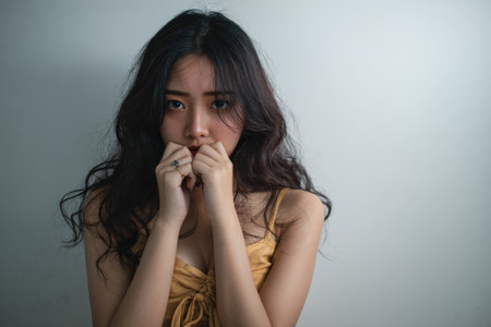 Young Asian woman expresses anxiety while covering her mouth with her hands in a minimalist indoor setting, reflecting distress and emotional turmoilの素材