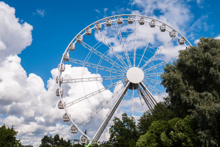 A large ferris wheel dominates the skyline, surrounded by lush greenery and bright blue skies filled with white clouds. People enjoy the vibrant park atmosphere nearby.の写真素材