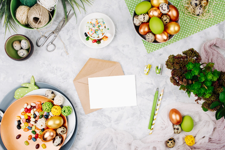 Workspace with easter decoration and empty card with envelope. Painted eggs, candy, flowers with copy space. Holiday background. Flat lay, top viewの写真素材