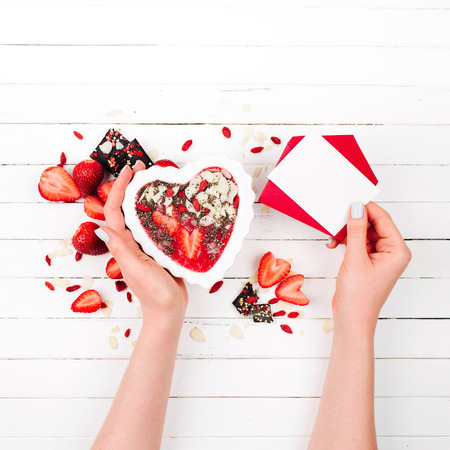 Female hands hold a strawberry smoothie in a bowl in the shape of a heart and valentine card. Valentine's Day. Holiday breakfastの写真素材