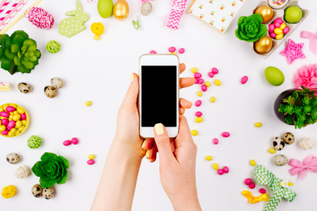 Female hand holding a smartphone.Beautiful spring mock up. Workspace with easter decoration. Flat lay, top viewの写真素材