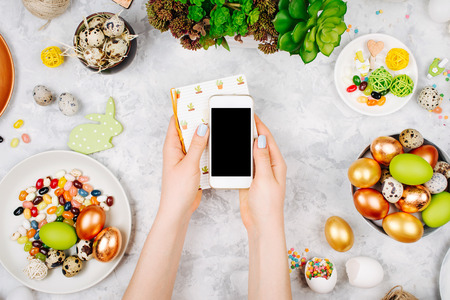 Female hand holding a notebook,  smartphone mock up.  Workspace with easter decoration. Painted eggs in trays, candy, flowers with copy space. Flat lay, top view の写真素材