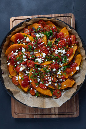 Delicious Hokkaido pumpkin with curry, mini tomatoes, hazelnuts, pumpkin seeds, dates and sheep's cheese baked on a dark rustic background, top view, verticalの写真素材