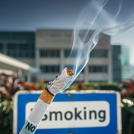Burning cigarette in front of a no smoking sign. A burning cigarette leans against a No Smoking sign with clear smoke, with a modern building under a blue sky in the background.の素材