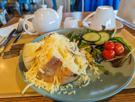 A wholesome meal featuring a generously cheese-topped baked potato alongside a crisp mixed salad, served in a bright restaurant setting with a pot of tea.の写真素材