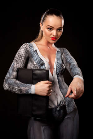 View of a sensual naked woman covered in body paint reperesenting office suit, holding a paper case and showing with her finger. High resolution concept image in studio on black backgroundの写真素材