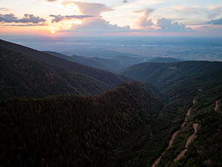 Aerial drone view of winding mountain road through forest valley at sunsetの写真素材