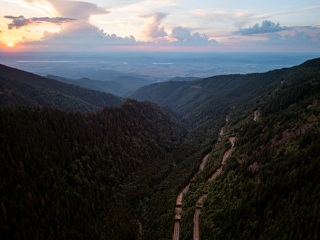 Aerial drone view of winding mountain road through forested valley at sunsetの写真素材