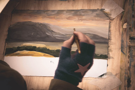 in the artists hand is a brush that paints a landscape on paper. watercolor painting