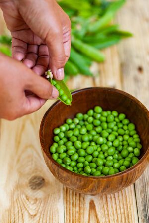 closeup of woman hand depod green peas in brown dish, on wooden table.の写真素材