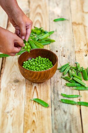 closeup of woman hand depod green peas in brown dish, on wooden table.の写真素材
