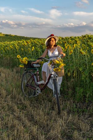 A woman in a hat and a white dress with a bicycle walks through the pollen with sunflowersの写真素材