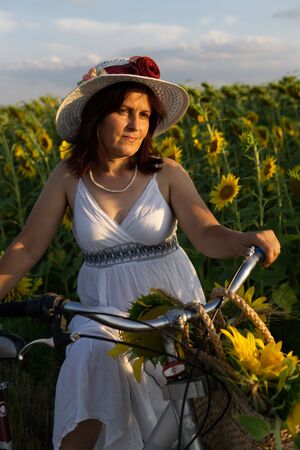 A woman in a hat and a white dress with a bicycle walks through the pollen with sunflowersの写真素材