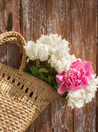 Flowers of pink red and white peonies in wicker basket on wooden table against wooden backgroundの写真素材