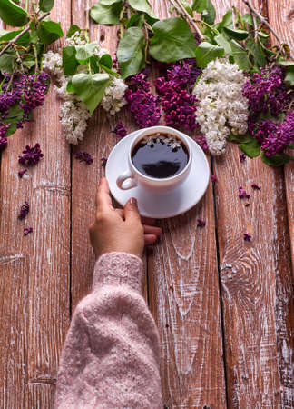 Hand holds a cup of morning coffee with spring lilac flowers branches blossoming on wooden background view from above. Flat lay underground style. Expensive colors. Creative design of flowers.の写真素材