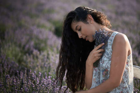 Beautiful girl in a white sarafan sits on a wooden chair and poses in the middle of a lavender field.の写真素材
