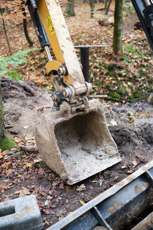 Close-up of an excavator bucket digging into the ground. A compact loader is parked carefully amidst colorful autumn leaves in a peaceful forest settingの写真素材