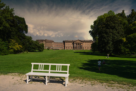 Kassel, Germany - 15 08 2024. An Elegant Bench Situated in a Scenic Park Surrounded by a Beautiful Historic Building Backdropの写真素材