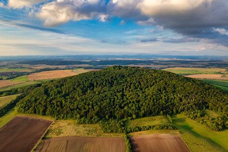 Aerial view of forested hill surrounded by patchwork farmland under dramatic clouds, lush canopy and rollingの写真素材