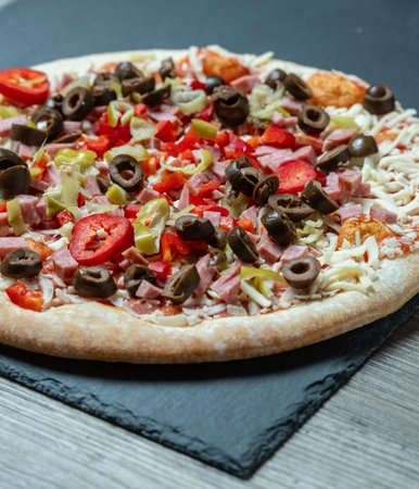 Overhead pizza with olives and tomatoes, full round pie on dark board beside gray wood surface,...