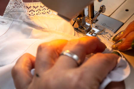 Close up view of sewing process. Female hands stitching white fabric on professional manufacturing machine at workplace. Seamstress hands holding textile for dress production. Light blurred backgroundの写真素材