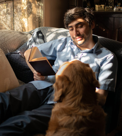 A relaxed man reading a book lying on a sofa next to a dog. Young smiling latin with black hair and...