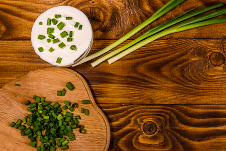 Glass bowl with sour cream and cutting board with chopped green onion on rustic wooden table. Top viewの写真素材