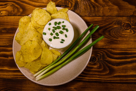 Ceramic plate with potato chips and glass bowl with sour cream on rustic wooden table. Top view