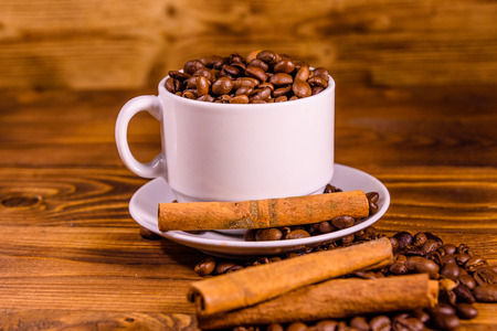 White cup filled with coffee beans and cinnamon sticks on rustic wooden tableの写真素材