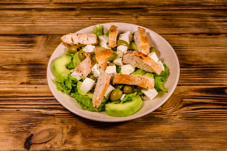 Fresh salad with chicken meat, feta cheese, avocado, green olives and lettuce leaves in ceramic plate on rustic wooden tableの写真素材