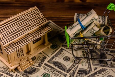Small shopping cart with rolled up one hundred dollar banknotes and plywood model of the house on wooden background. Loan, real estate conceptの写真素材