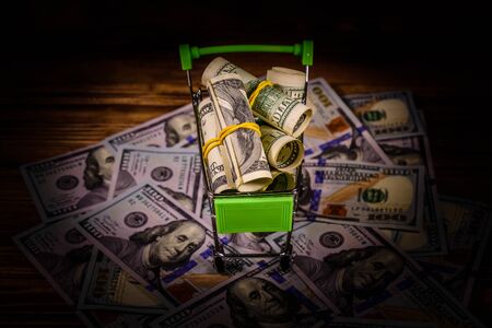 Small shopping cart with rolled up one hundred dollar banknotes on wooden backgroundの写真素材