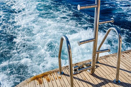 Metal staircase for descent into the water and wave trace with white foam on water surface behind of fast moving yachtの写真素材