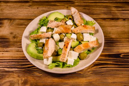 Fresh salad with chicken meat, feta cheese, avocado, green olives and lettuce leaves in ceramic plate on rustic wooden tableの写真素材