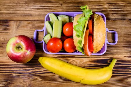 Ripe apple, banana and lunch box with hamburger, cucumbers and tomatoes on rustic wooden table. Top viewの写真素材
