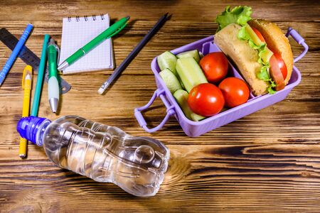Bottle of water, different stationeries and lunch box with hamburger, cucumbers and tomatoes on rustic wooden tableの写真素材