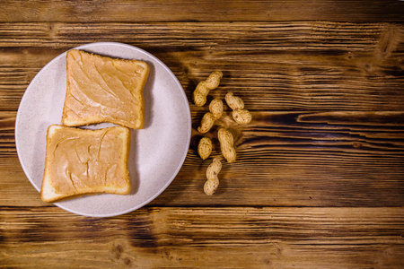 Glass jar with peanut butter and plate with sandwiches on wooden table. Top view