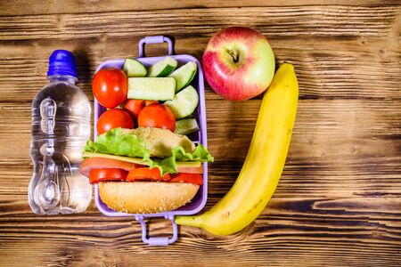 Ripe apple, banana, bottle of water and lunch box with hamburger, cucumbers and tomatoes on rustic wooden table. Top viewの写真素材