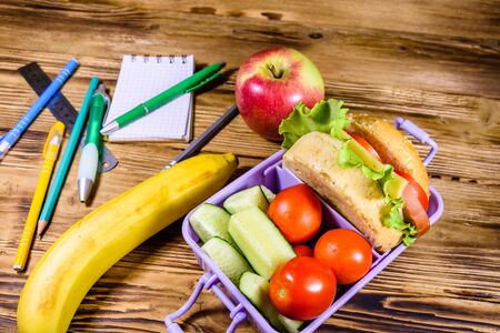 Ripe apple, banana, different stationeries and lunch box with hamburger, cucumbers and tomatoes on rustic wooden tableの写真素材